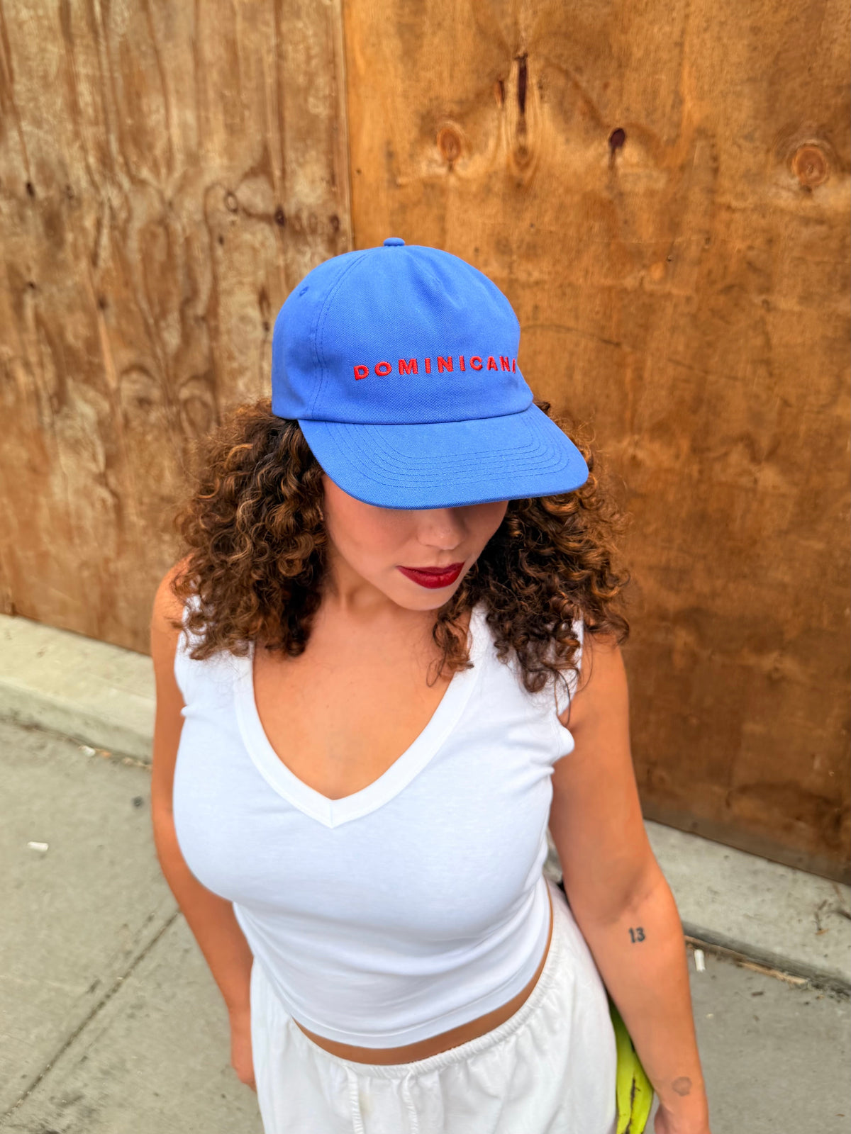 A woman wearing a bright blue baseball cap with 'DOMINICANA' embroidered in red across the front, paired with a white tank top and white skirt, standing outdoors against a wooden wall.