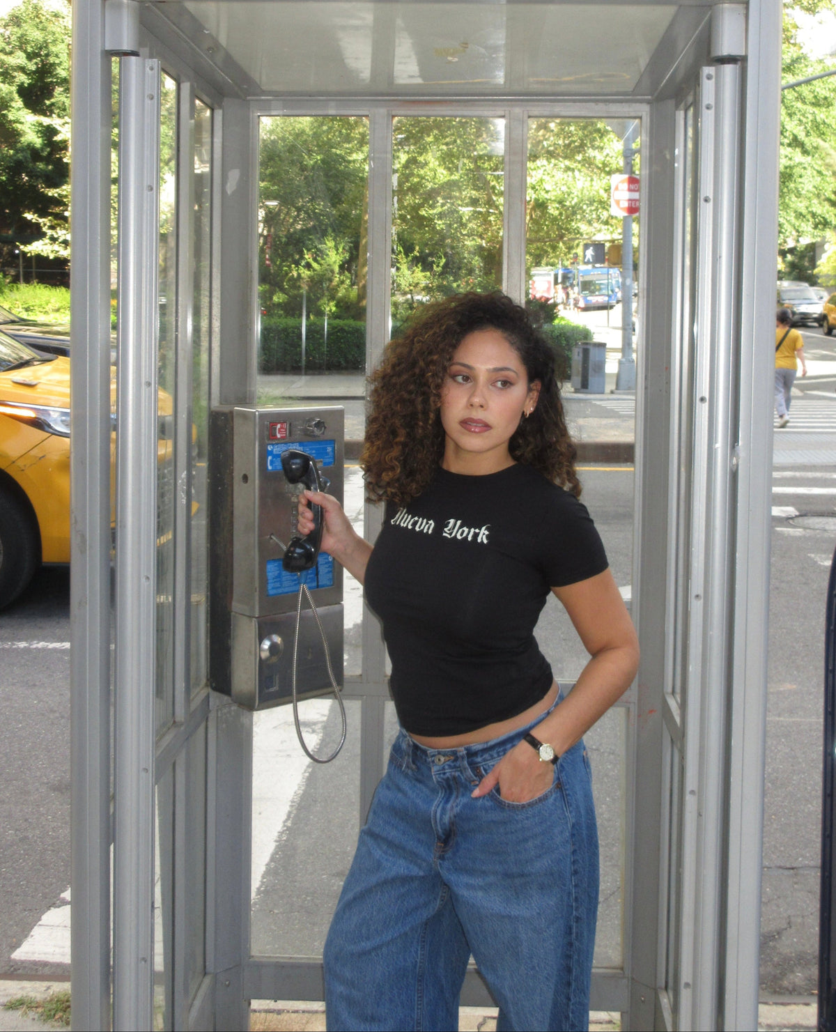 A woman with curly hair stands inside a silver public phone booth on a street, holding a blue payphone. She is wearing a black cropped t-shirt with 'New York' printed on it and blue jeans.