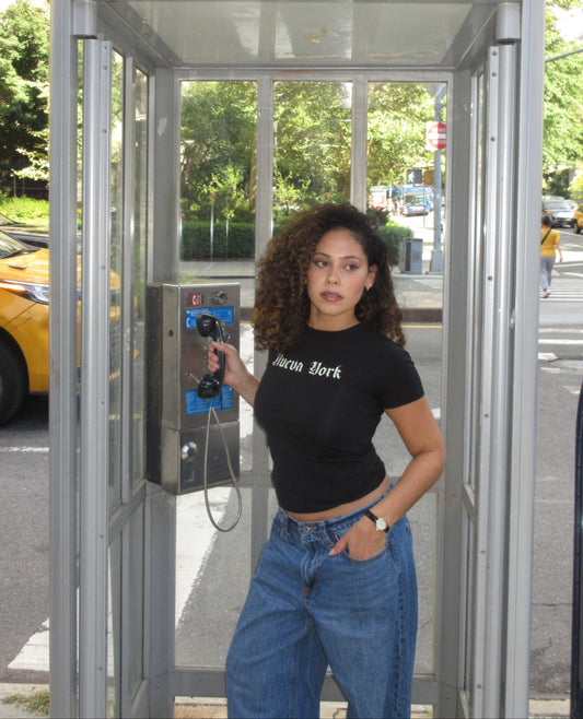 A woman with curly hair stands inside a silver public phone booth on a street, holding a blue payphone. She is wearing a black cropped t-shirt with 'New York' printed on it and blue jeans.